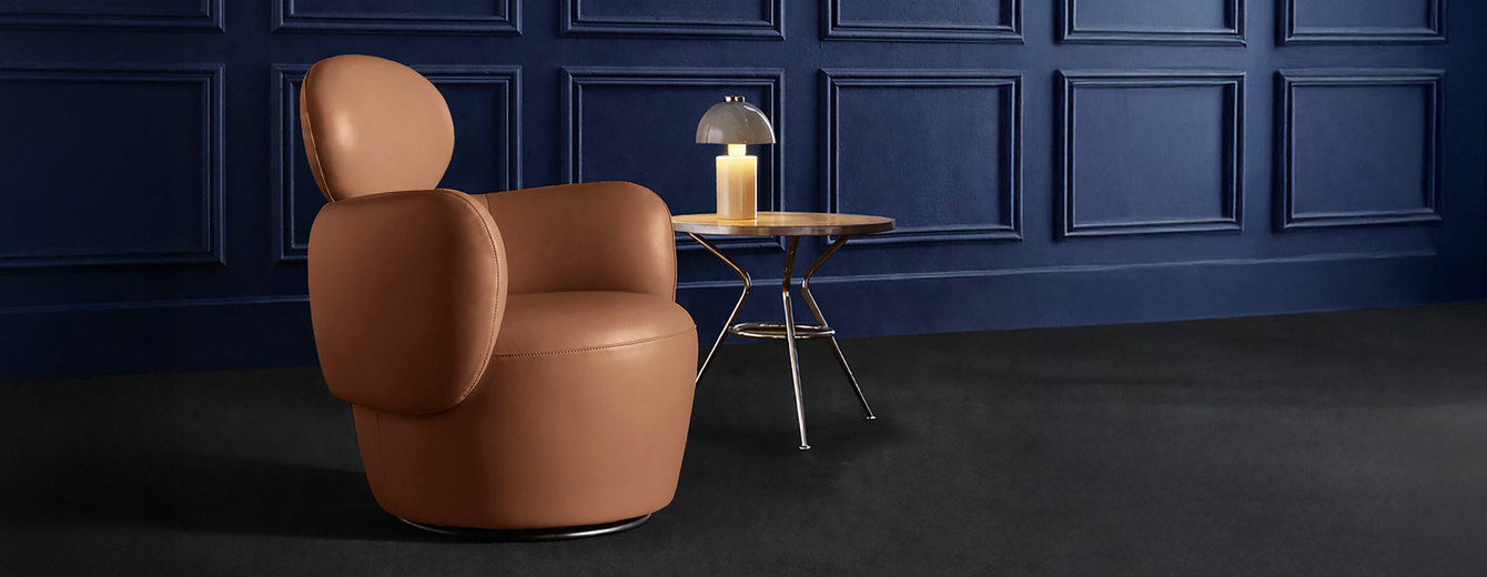 Fauteuil design en tissu beige aux formes arrondies, posé sur un sol rose poudré devant un mur bordeaux, dans une composition minimaliste et élégante.