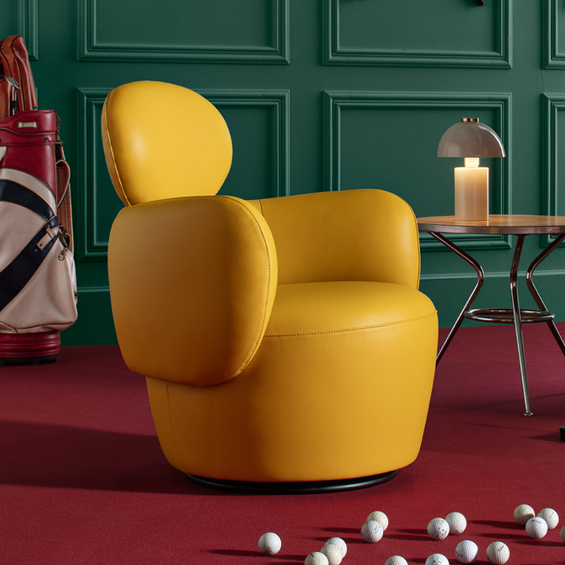 Yellow armchair with rounded design placed on a red carpet, in front of a green paneled wall, with a designer lamp and golf balls scattered on the floor.
