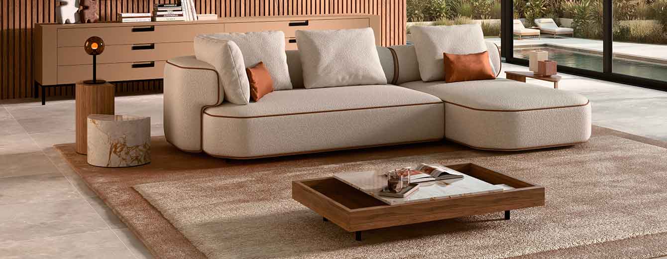 Curved contemporary sectional sofa upholstered in textured off-white fabric with warm leather piping, set in a modern living room featuring wooden slat walls, abstract art, and large floor-to-ceiling windows opening onto a landscaped terrace.