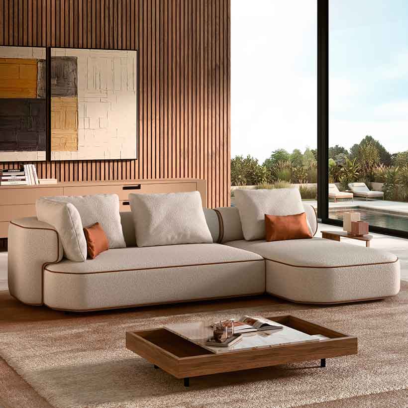 Curved contemporary sectional sofa upholstered in textured off-white fabric with warm leather piping, set in a modern living room featuring wooden slat walls, abstract art, and large floor-to-ceiling windows opening onto a landscaped terrace.