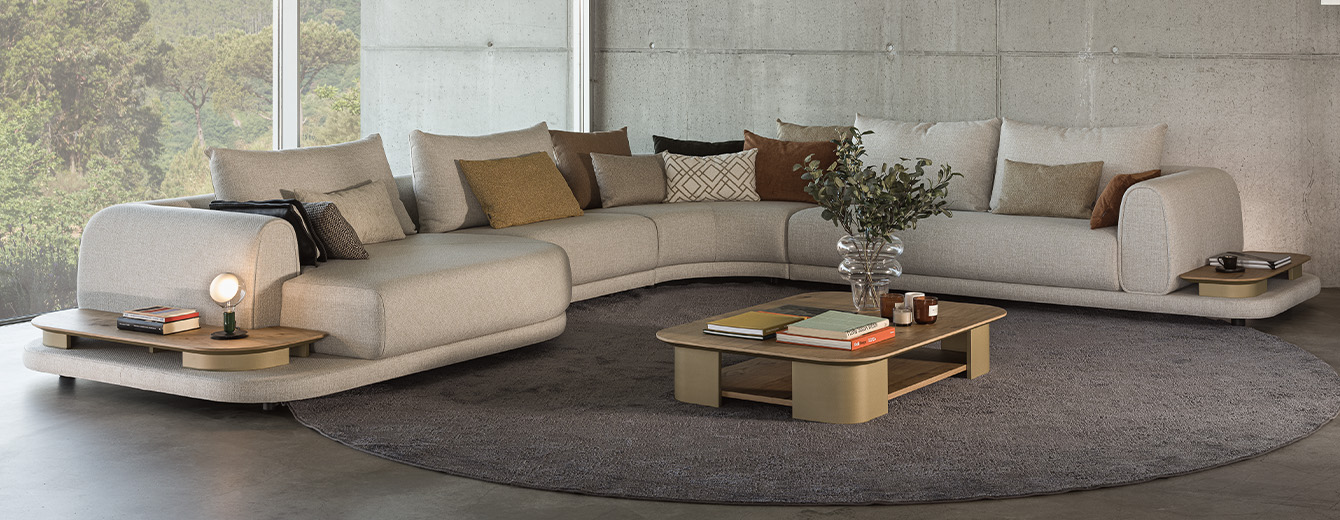 Contemporary living room with a beige sectional sofa and two green armchairs, arranged around oval coffee tables on a large gray rug, facing a picture window overlooking nature.