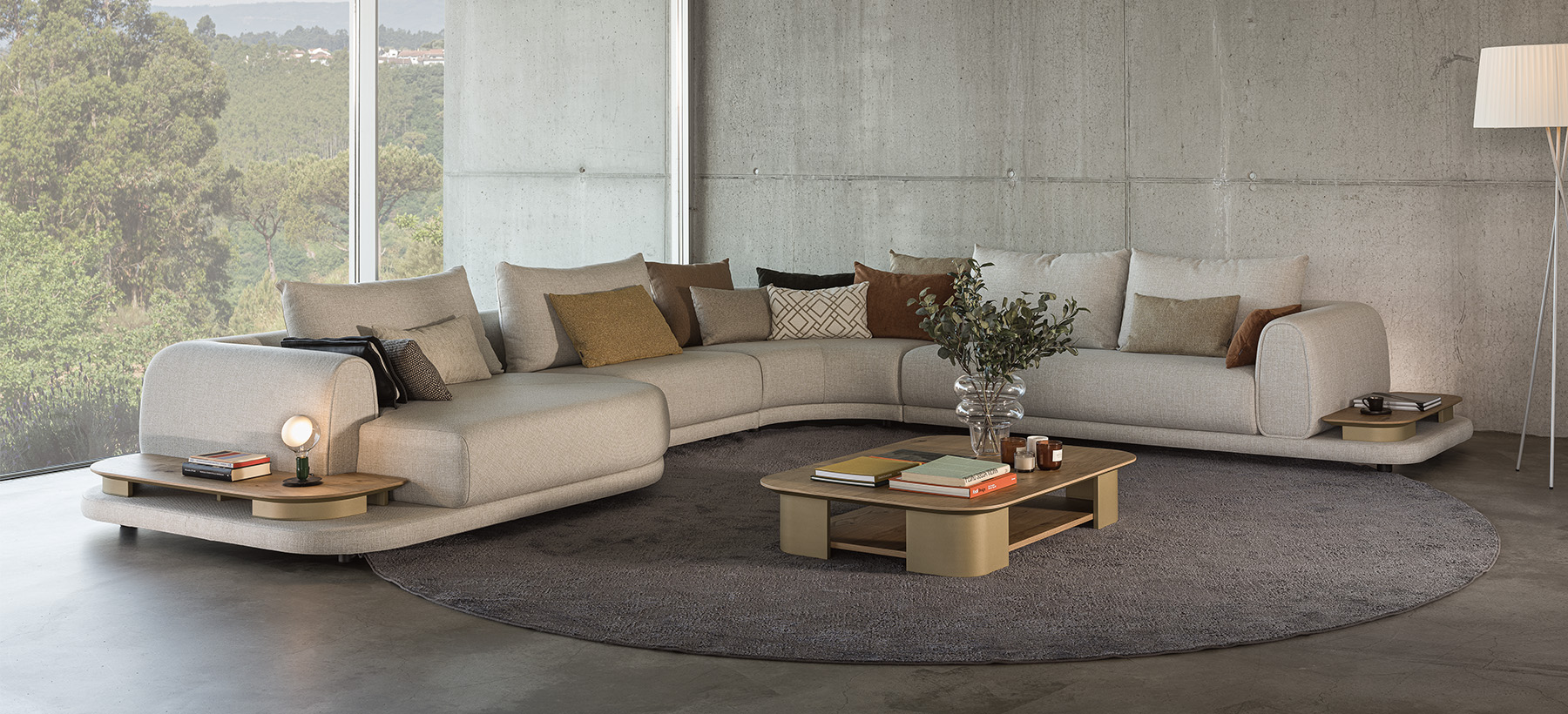 Contemporary living room with a beige sectional sofa and two green armchairs, arranged around oval coffee tables on a large gray rug, facing a picture window overlooking nature.