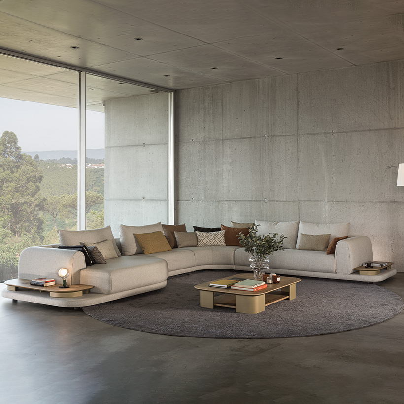 Salon contemporain avec canapé d’angle beige et deux fauteuils verts, disposés autour de tables basses ovales sur un grand tapis gris, face à une baie vitrée ouverte sur la nature.