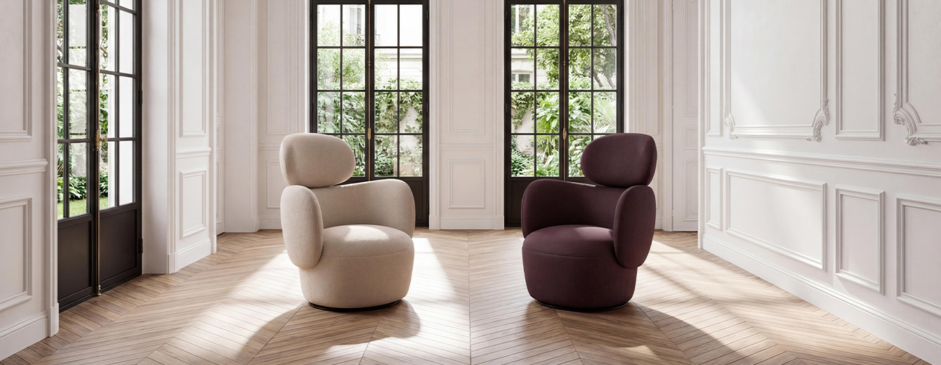 Two fabric armchairs with rounded shapes, one beige and one burgundy, set on a herringbone parquet floor in a bright Parisian interior with white moldings and large windows overlooking lush greenery.